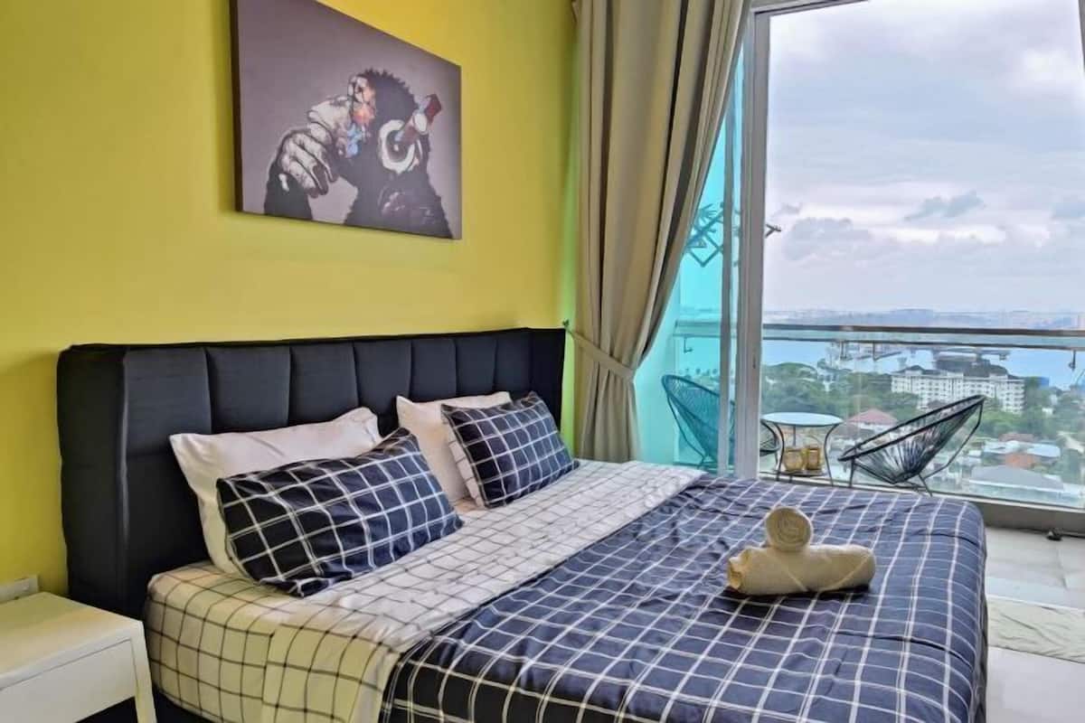 Une chambre à coucher avec un lit, des oreillers, un tableau, une fenêtre donnant sur un paysage et une chaise.