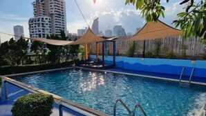 Piscina con un paisaje urbano al fondo, rodeada de vegetación y zonas con sombra.