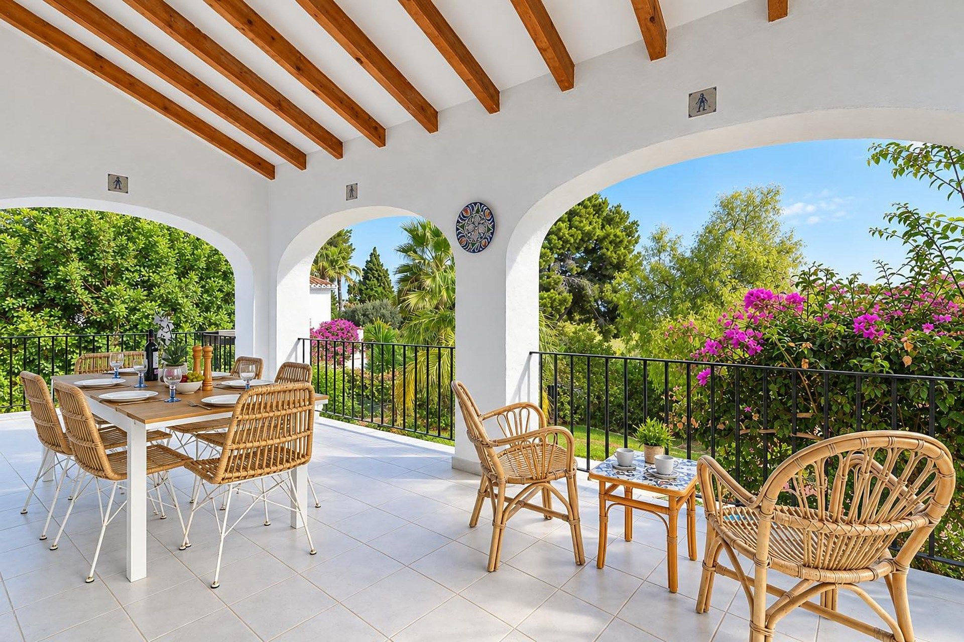 An outdoor patio with a table set for dining, surrounded by wicker chairs, under a white ceiling with wooden beams.