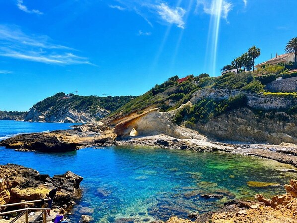 A coastal landscape with clear waters, rocky terrain, and a distant hill with vegetation.
