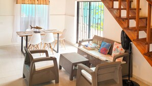Amplia sala de estar con techo alto, escalera de madera, mobiliario de mimbre y una mesa de comedor con sillas.