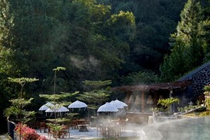 An outdoor dining area with tables and umbrellas, surrounded by lush greenery and a water feature. - Joglo Ageng Guci Tegal (Bumijawa)