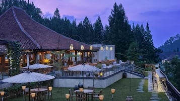 Un área de comedor al aire libre con mesas y sombrillas, un edificio con techo de tejas y un fondo de bosque.