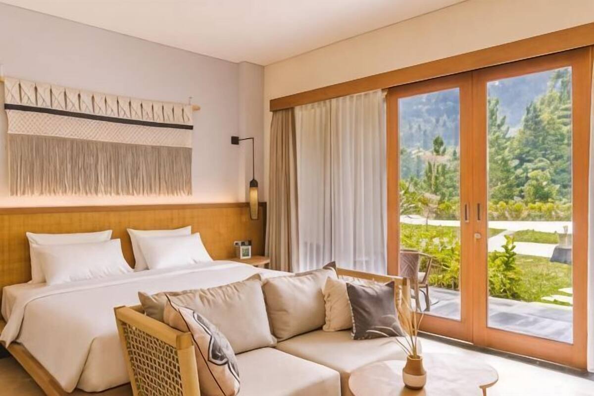 A hotel room with a bed, a sofa, a table, and a view of greenery.