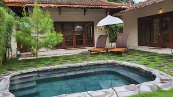 Una piscina con bordes de piedra, un patio cubierto con sillas y una estructura de techo de paja.