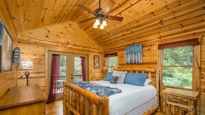 Ein Schlafzimmer einer Holzhütte mit Bett, Deckenventilator und Fenster mit Blick auf den Wald.