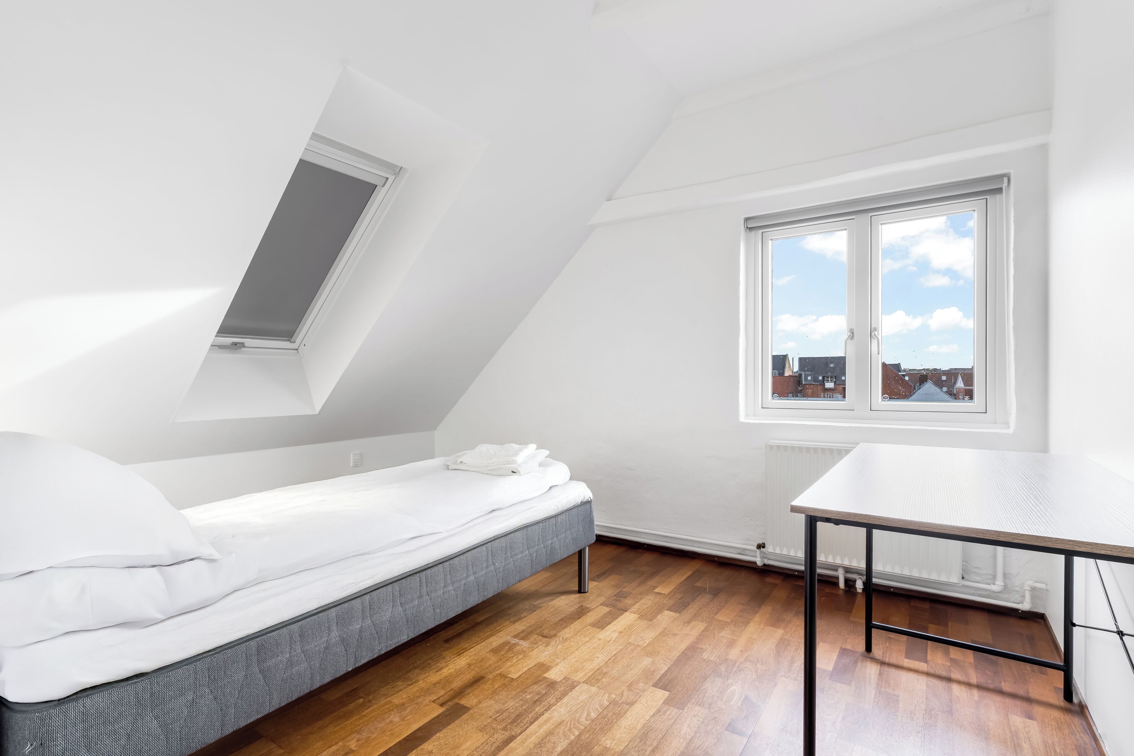 A small, bright room with a sloped ceiling, a single bed, a table, and a window with a view of buildings.