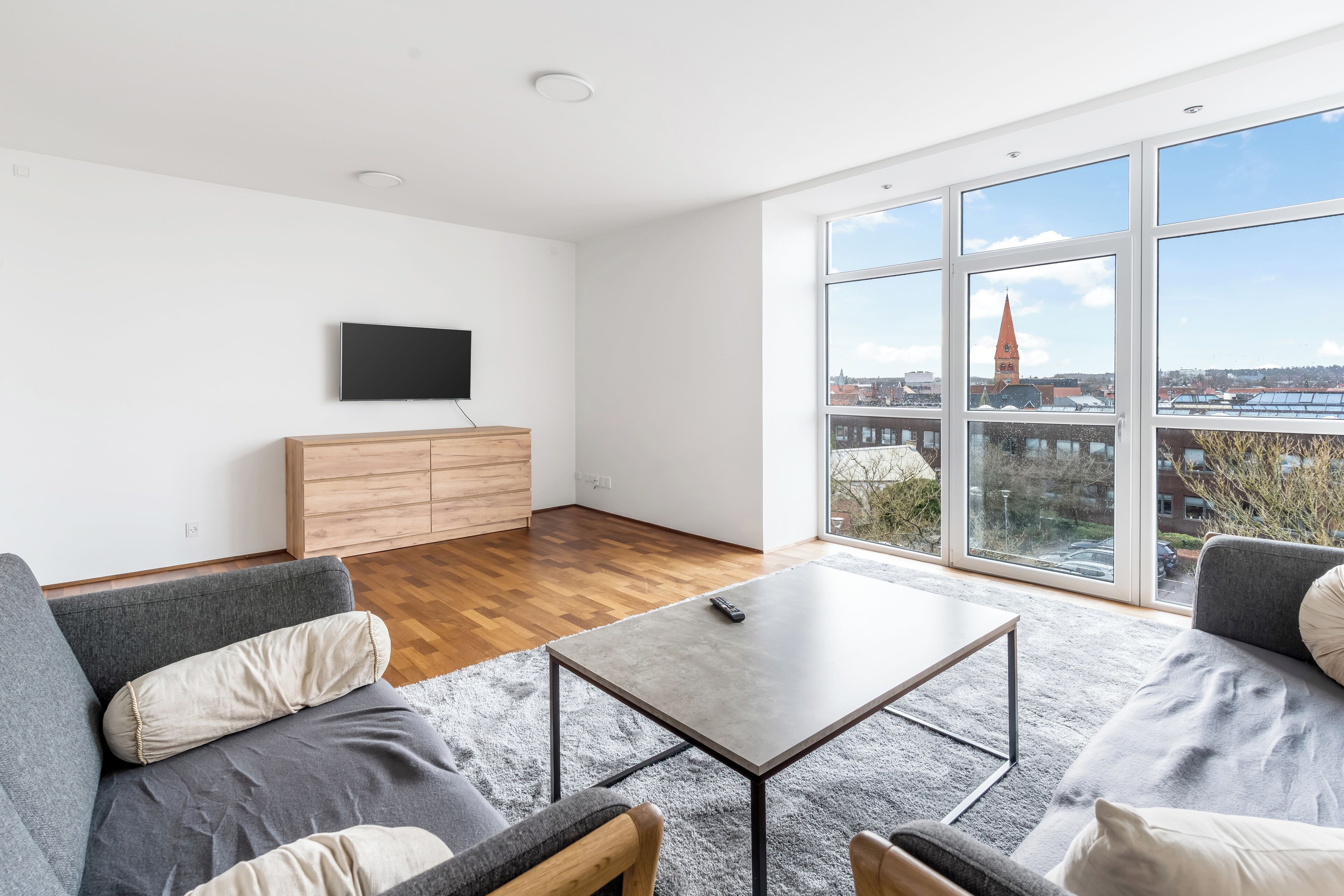 A modern living room with a flat-screen TV, wooden furniture, a coffee table, and large windows offering a city view.