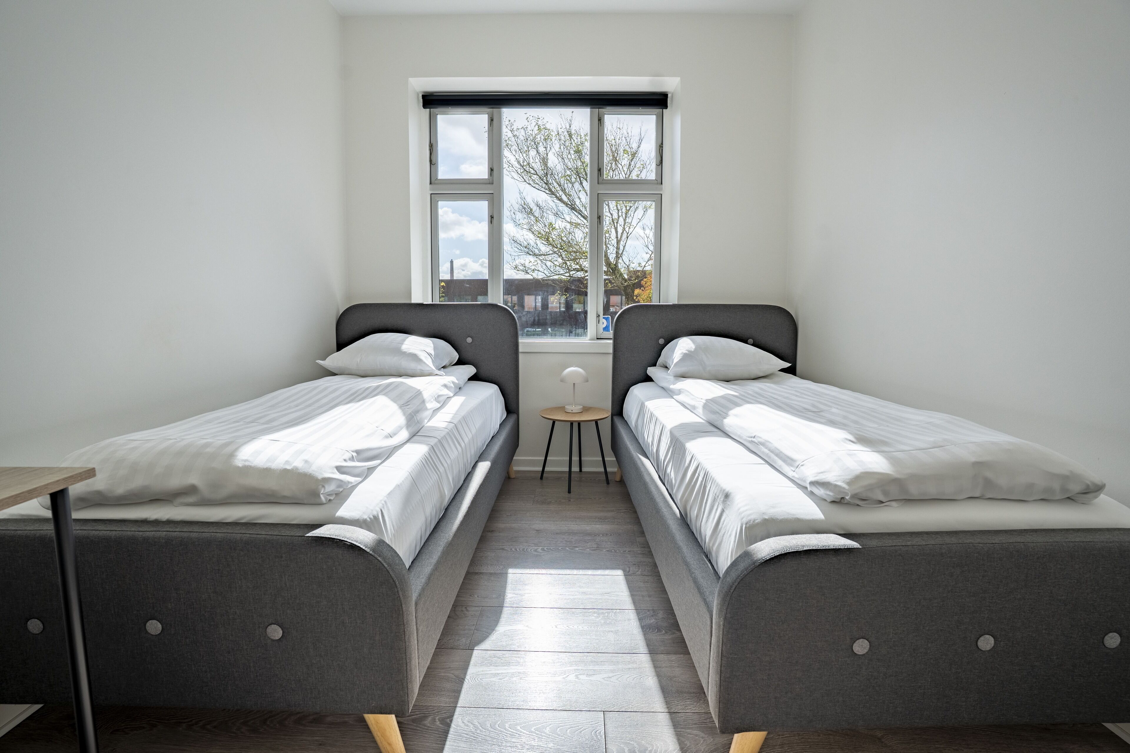 A bedroom with two single beds, a small table, and a window with a view of buildings and trees.