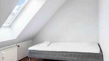 Une petite chambre avec un lit simple, un plafond en pente et une fenêtre laissant entrer la lumière naturelle.