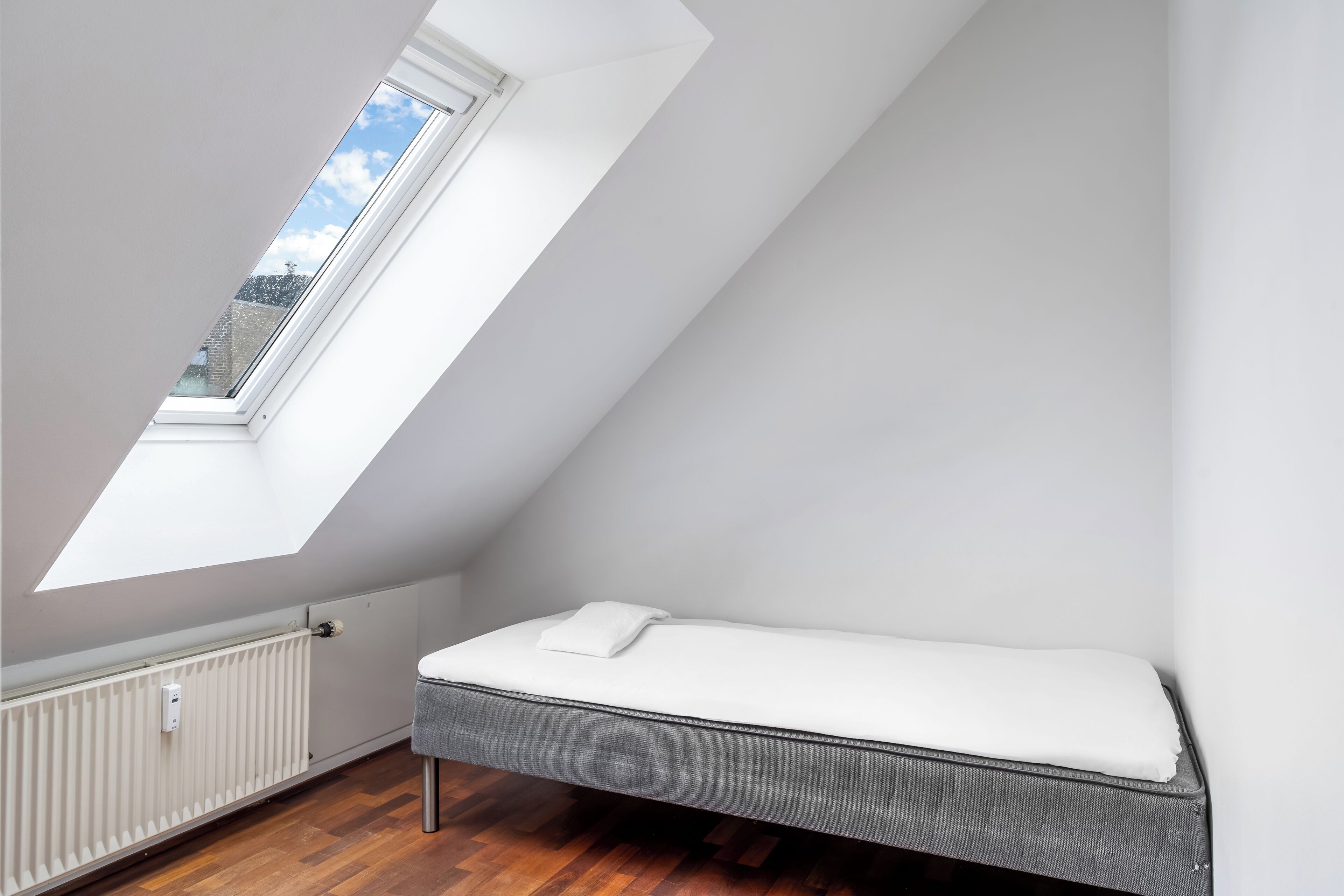 Una habitación pequeña con techo inclinado, tragaluz, cama y radiador.