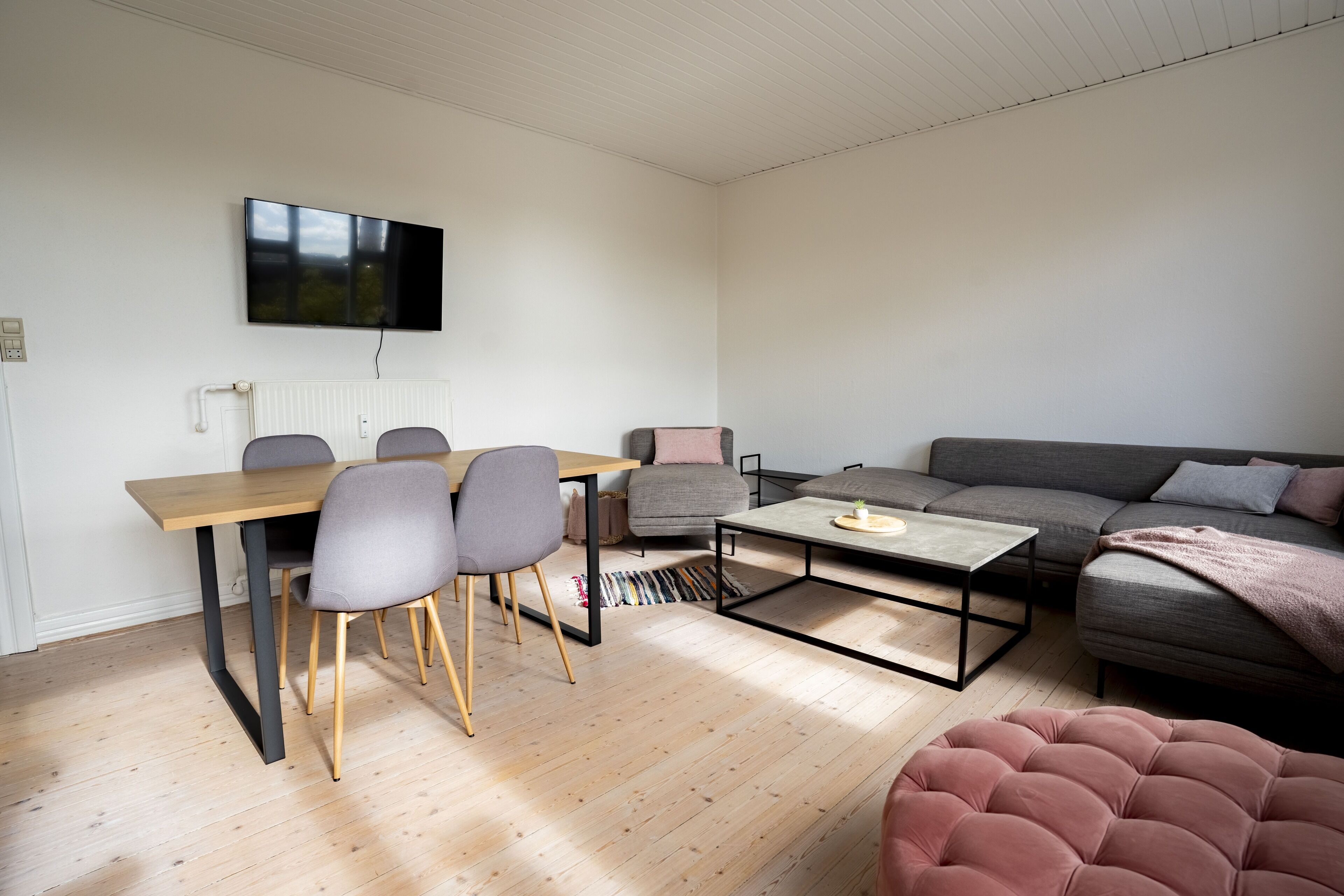 A modern living room with a wooden dining table, grey chairs, a grey sofa, and a pink ottoman.