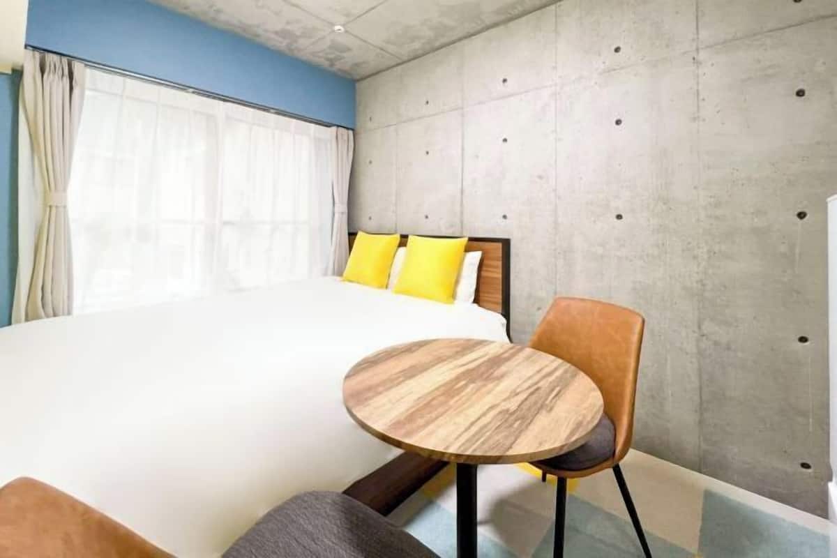 Un dormitorio con una cama, una mesa redonda de madera, una silla y una pared de concreto como detalle decorativo.