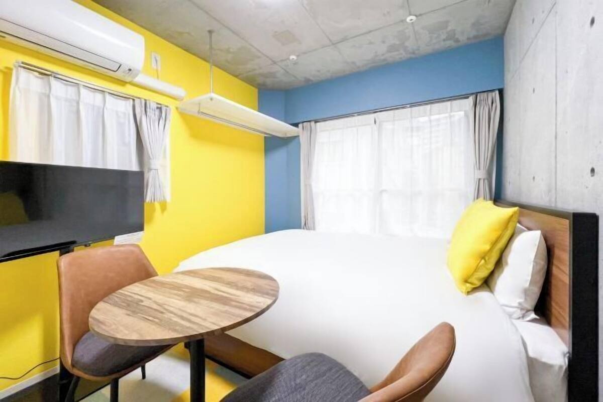 A hotel room with a bed, a small round table, a chair, a TV, and an air conditioning unit.