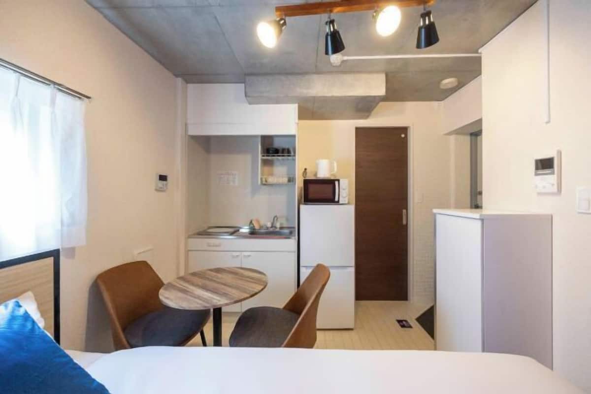 Habitación de hotel compacta con cama, una pequeña mesa de comedor con sillas, kitchenette y zona de ducha.