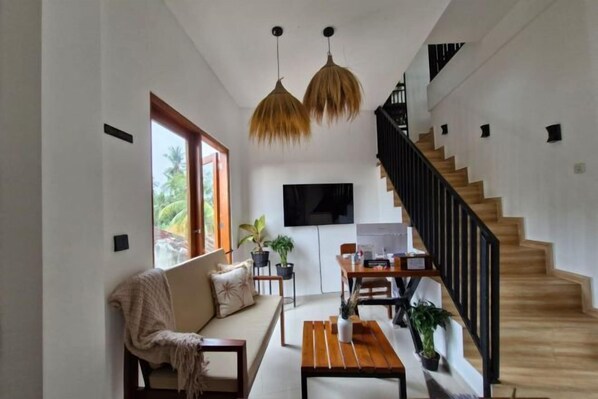 A spacious living area with a sofa, coffee table, and dining table, a staircase, and a TV mounted on the wall.