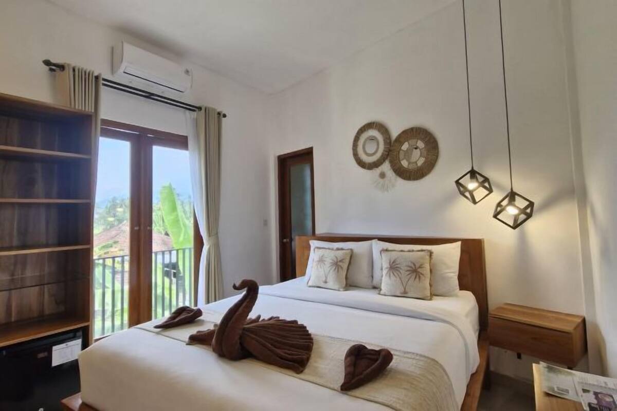 A hotel room with a bed, a wooden nightstand, a wall-mounted air conditioner, and a balcony with a view.