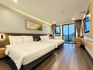A hotel room with two beds, a large window with curtains, a mounted artwork, and a small desk area.