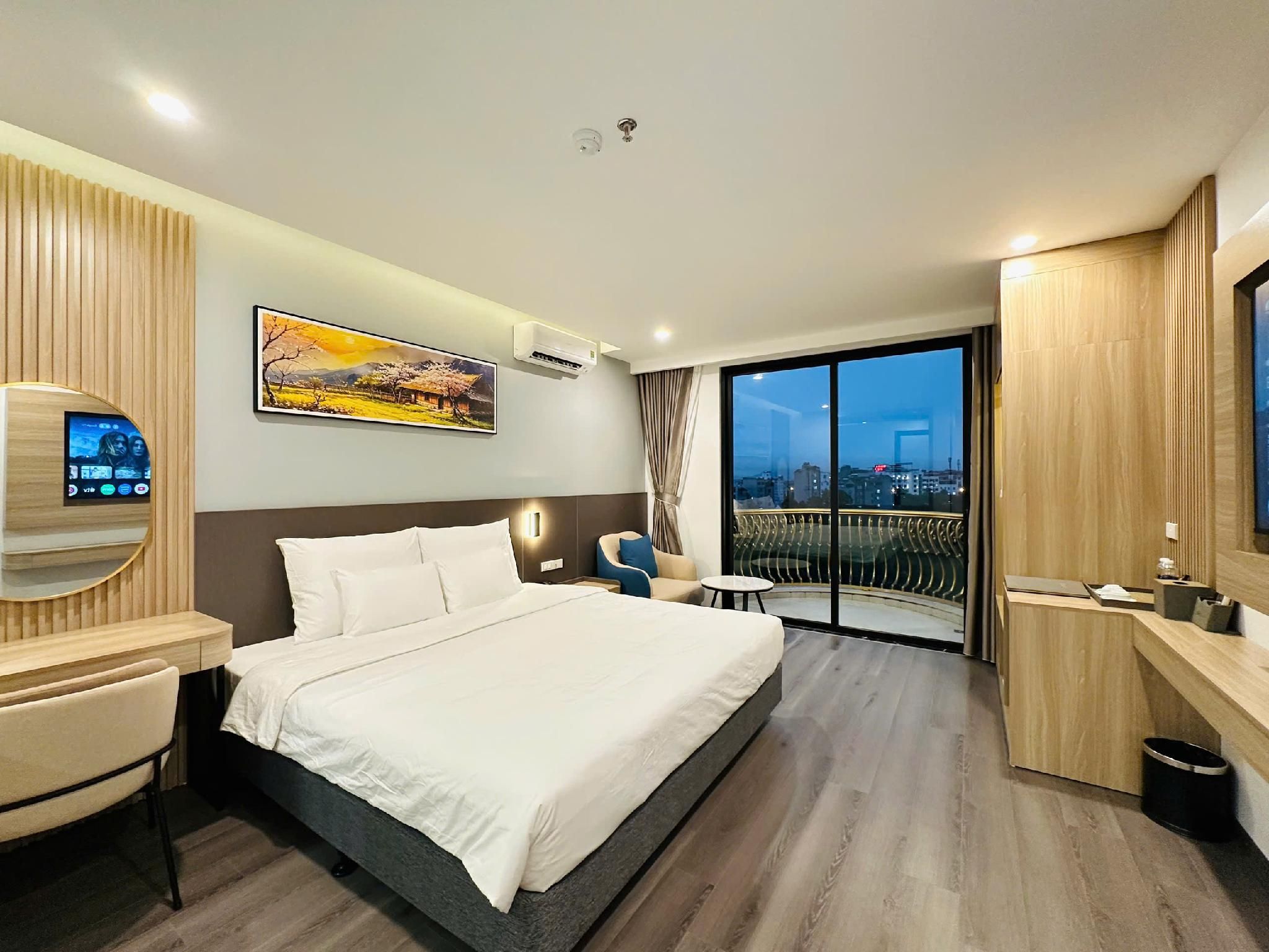 A modern hotel room with a large bed, a desk, a chair, a TV, and a balcony with a city view.