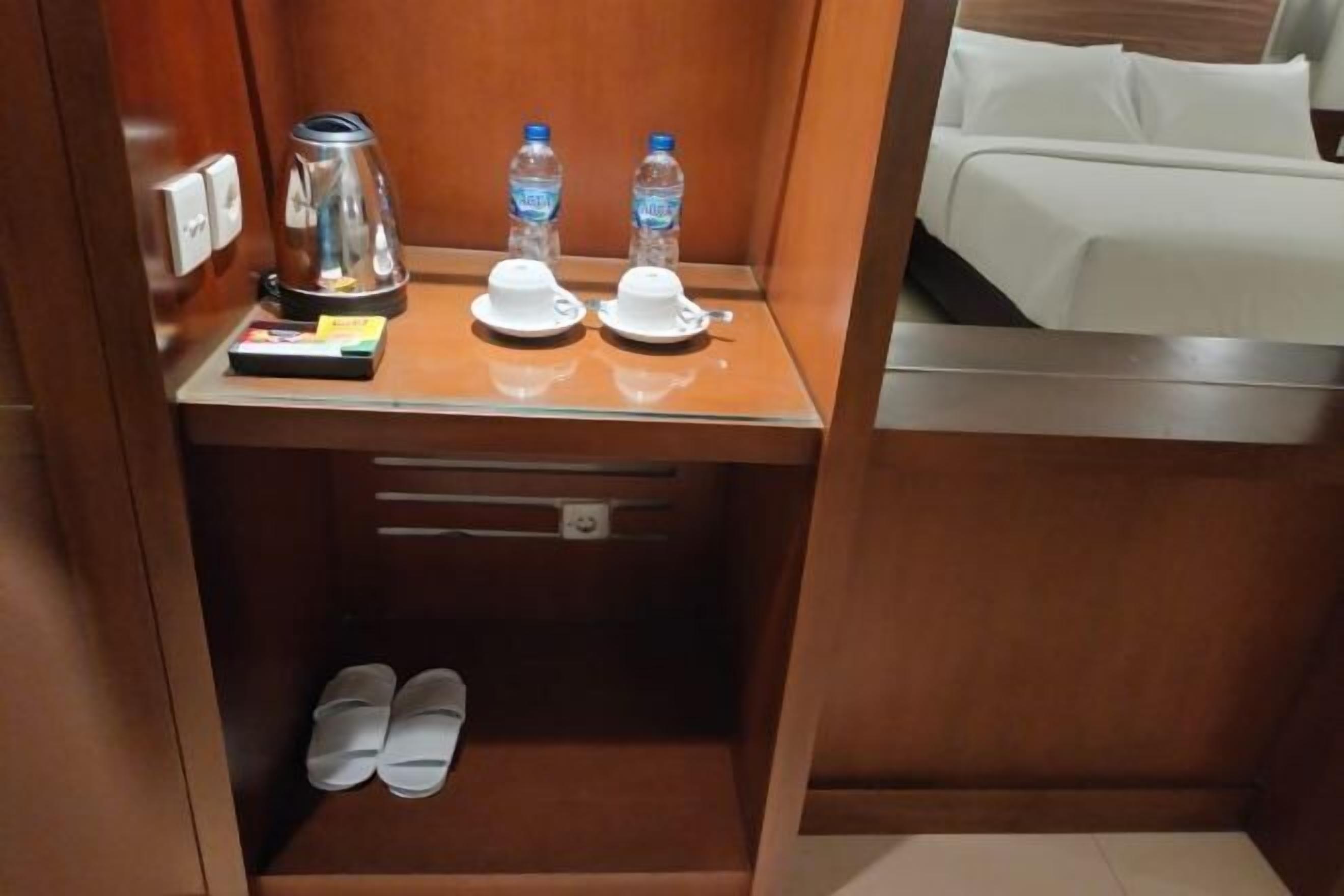A compact hotel room with a wooden nightstand, a kettle, two water bottles, a pair of slippers, and a bed with white bedding.