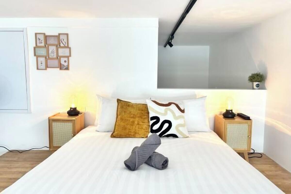Una cama bien hecha con una toalla, dos mesitas de noche con lámparas, un estante en la pared con una planta y una colección de cuadros enmarcados.