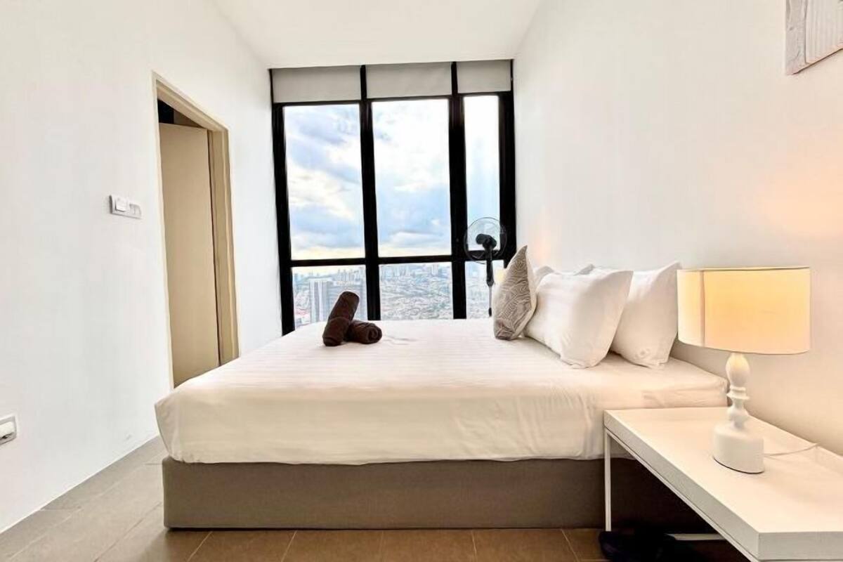 Una habitación de hotel moderna con un ventanal grande, una cama con sábanas blancas, una mesita de noche con una lámpara y vistas a la ciudad.