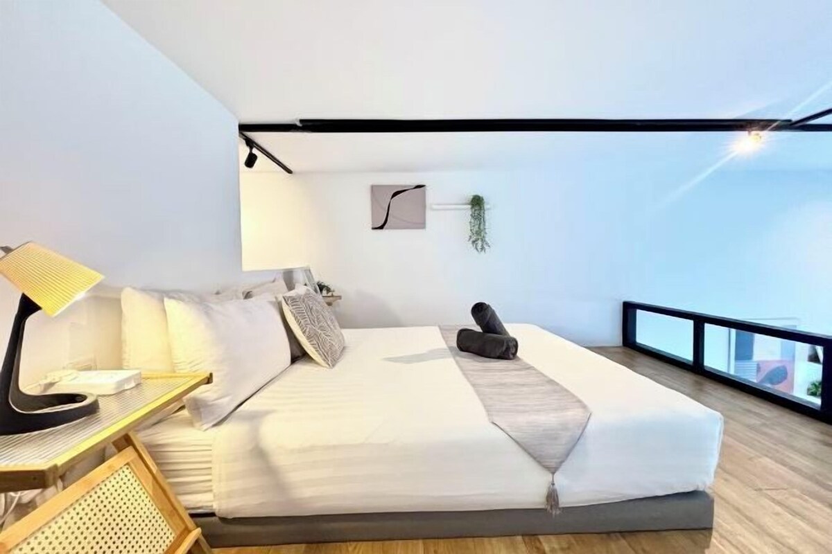 A modern hotel room with a large bed, a desk with a lamp, a small table, and a view of the city through a balcony railing.