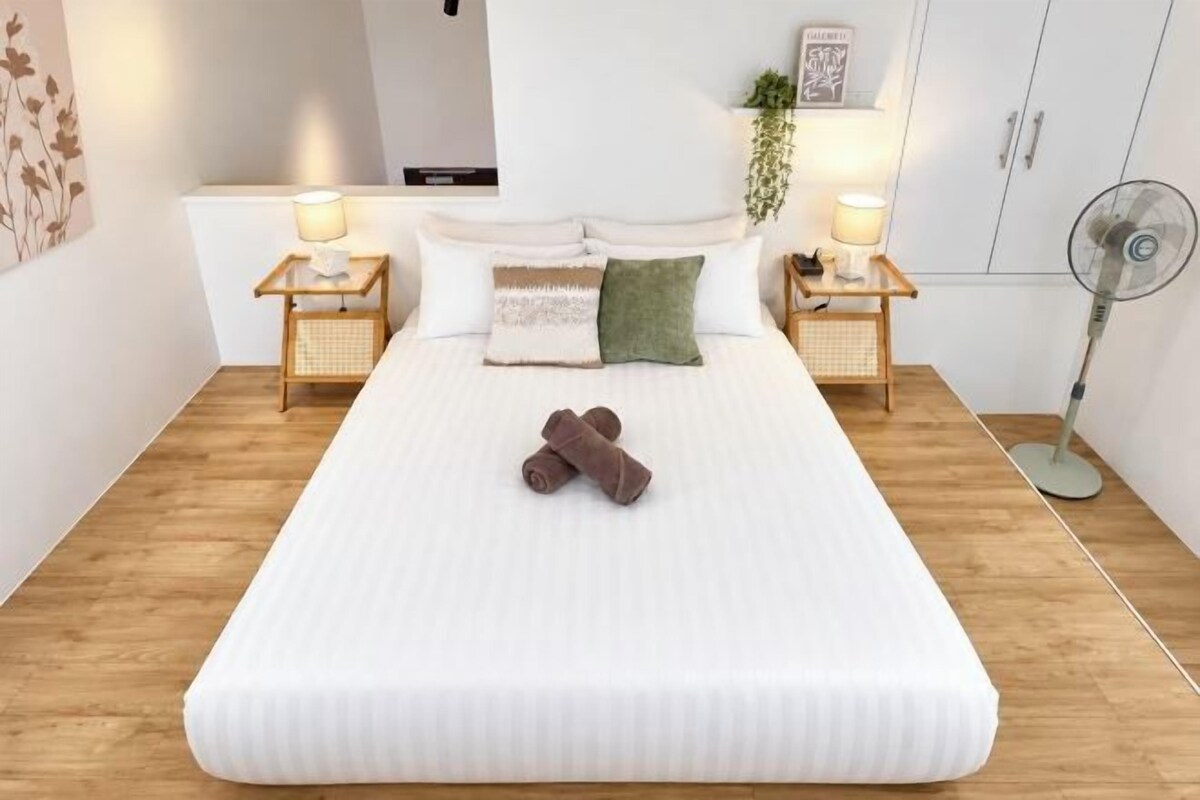 A neatly made bed with pillows and a blanket, two bedside tables with lamps, a wall-mounted fan, and a plant on the wall.