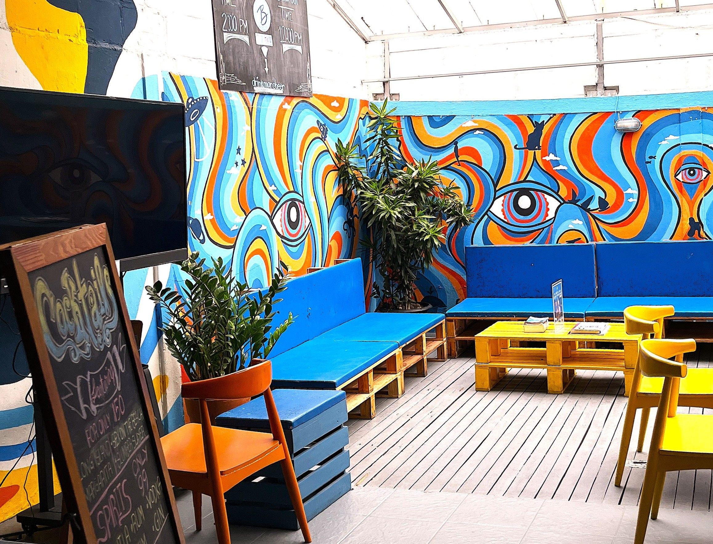 Un área de estar al aire libre con colores vivos, un mural, muebles de madera y un menú en una pizarra.