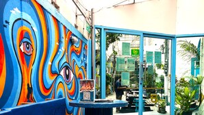 Une fresque colorée ornée d’un motif représentant un œil, un banc bleu et une porte vitrée donnant accès à un restaurant.