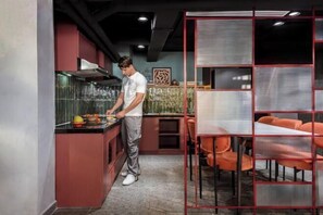 Una cocina moderna con una persona cocinando, decoración en rojo y negro, y sillas naranjas en una zona de comedor.