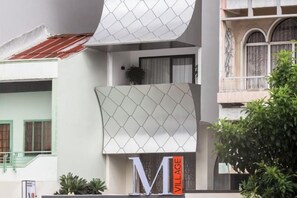 Un edificio moderno con un llamativo letrero 'M VILLAGE', un balcón con una planta en maceta y una ventana tradicional con celosía decorativa.