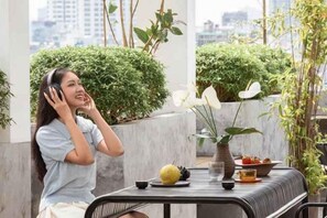 A person sitting at an outdoor table with a beverage and food items, surrounded by greenery and a cityscape view. - Express by M Village Vo Van Kiet, a brand of Modern Village Lifestyle (Ho Chi Minh City)