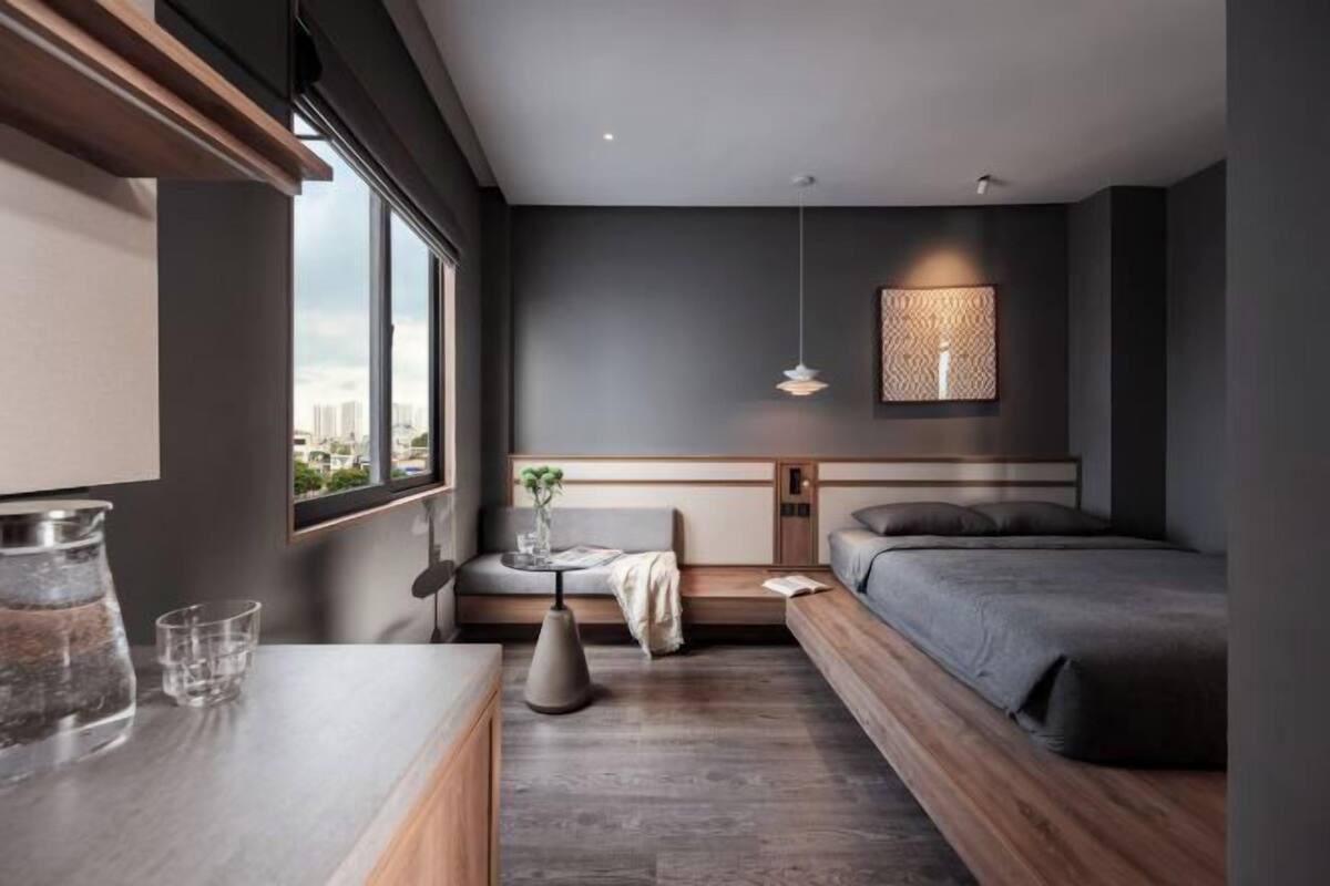 A modern bedroom with a large window, a bedside table with a vase and glasses, a small round table, and a bed with a headboard.