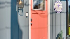 A door with a red frame and a window with blinds, a light fixture above it, and a circular sign with a bird and text. - 'Blue Heron' Walk to Kelso Beach! Pet-Friendly Gem (Erie)