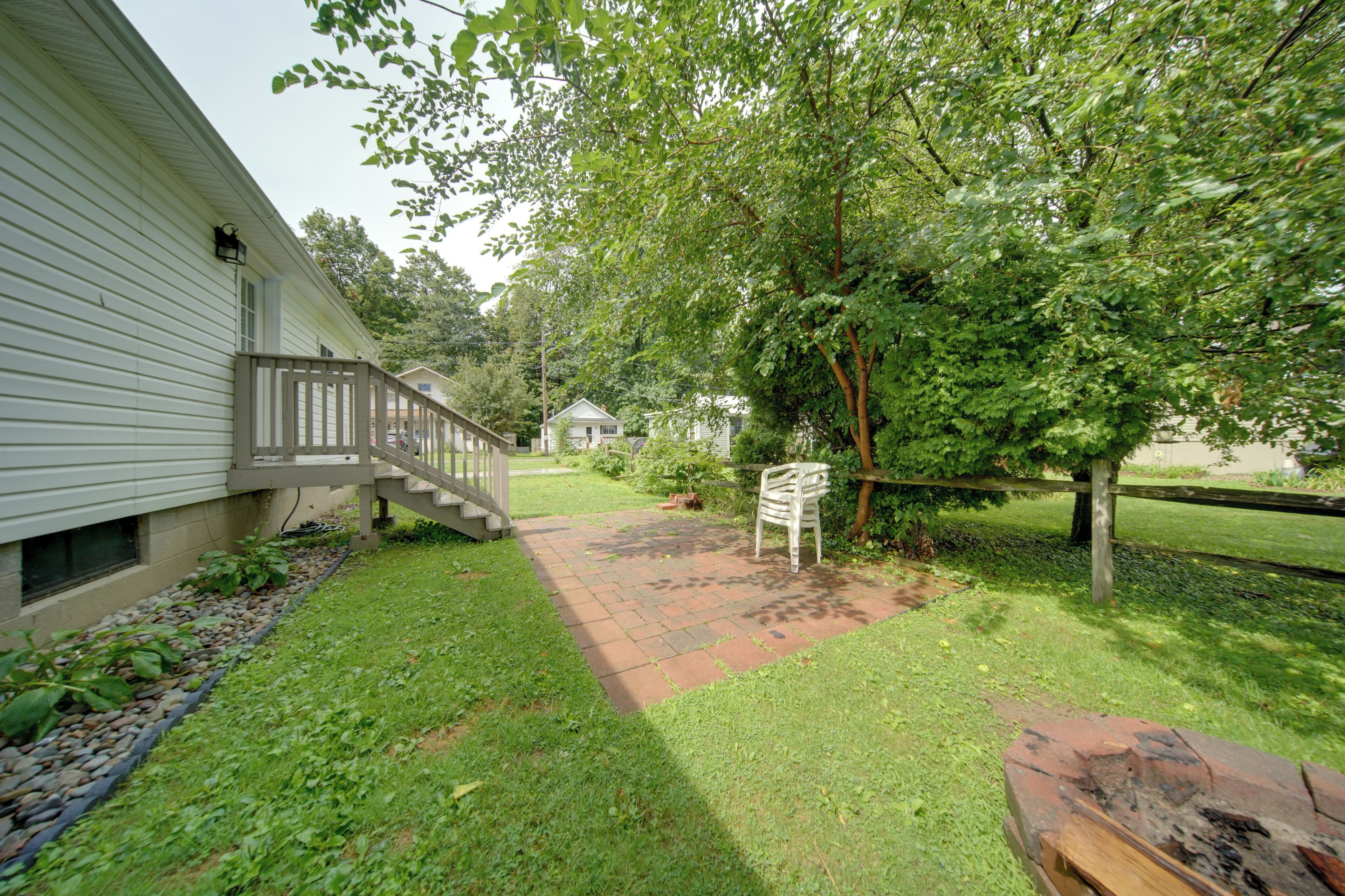 A residential property with a paved pathway leading to a wooden deck, a small house in the background, and a large tree providing shade.