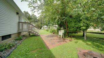 A residential property with a paved pathway leading to a wooden deck, a small house in the background, and a large tree providing shade.