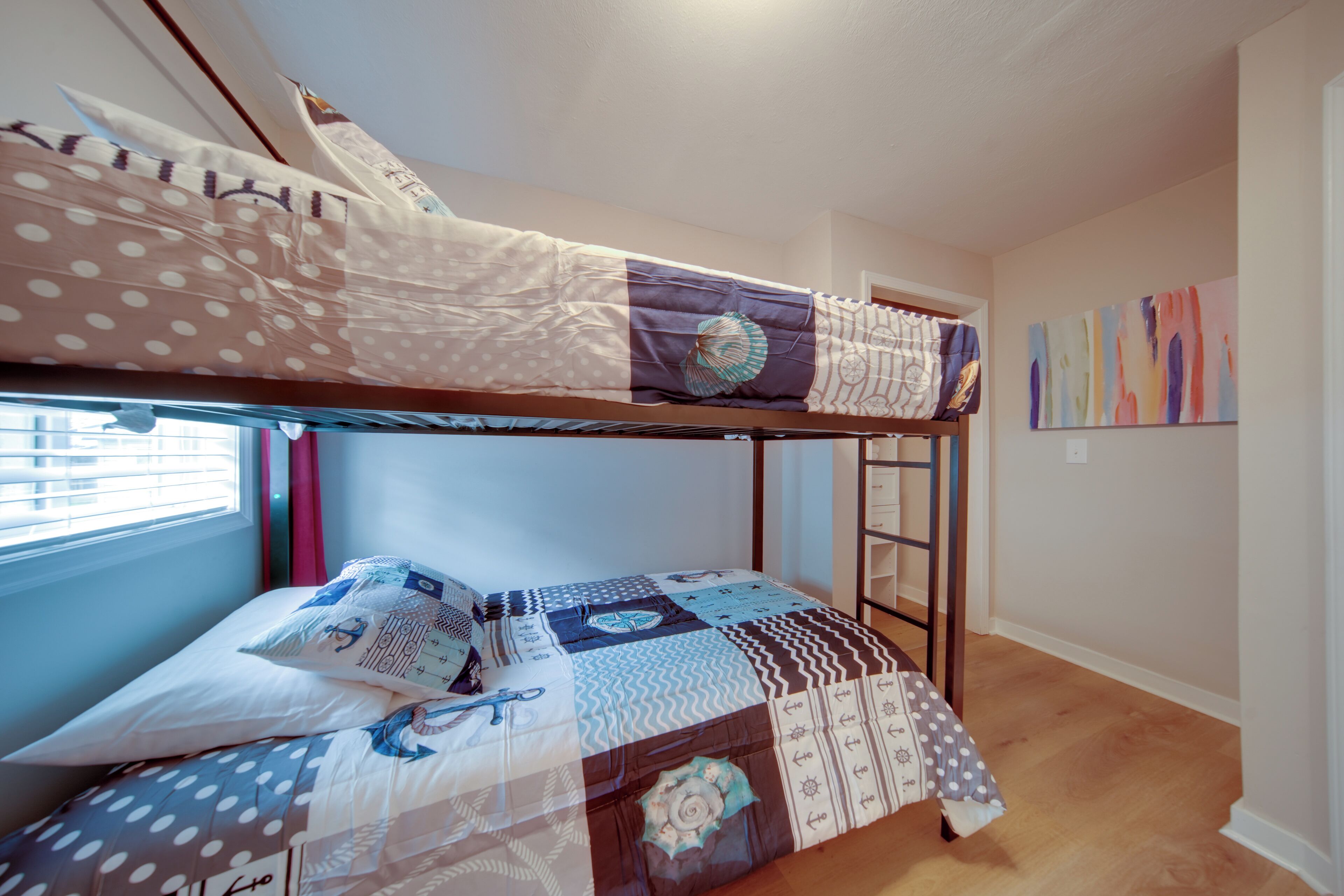 A bunk bed with a polka dot and striped duvet, a window with blinds, and a wall with abstract art.