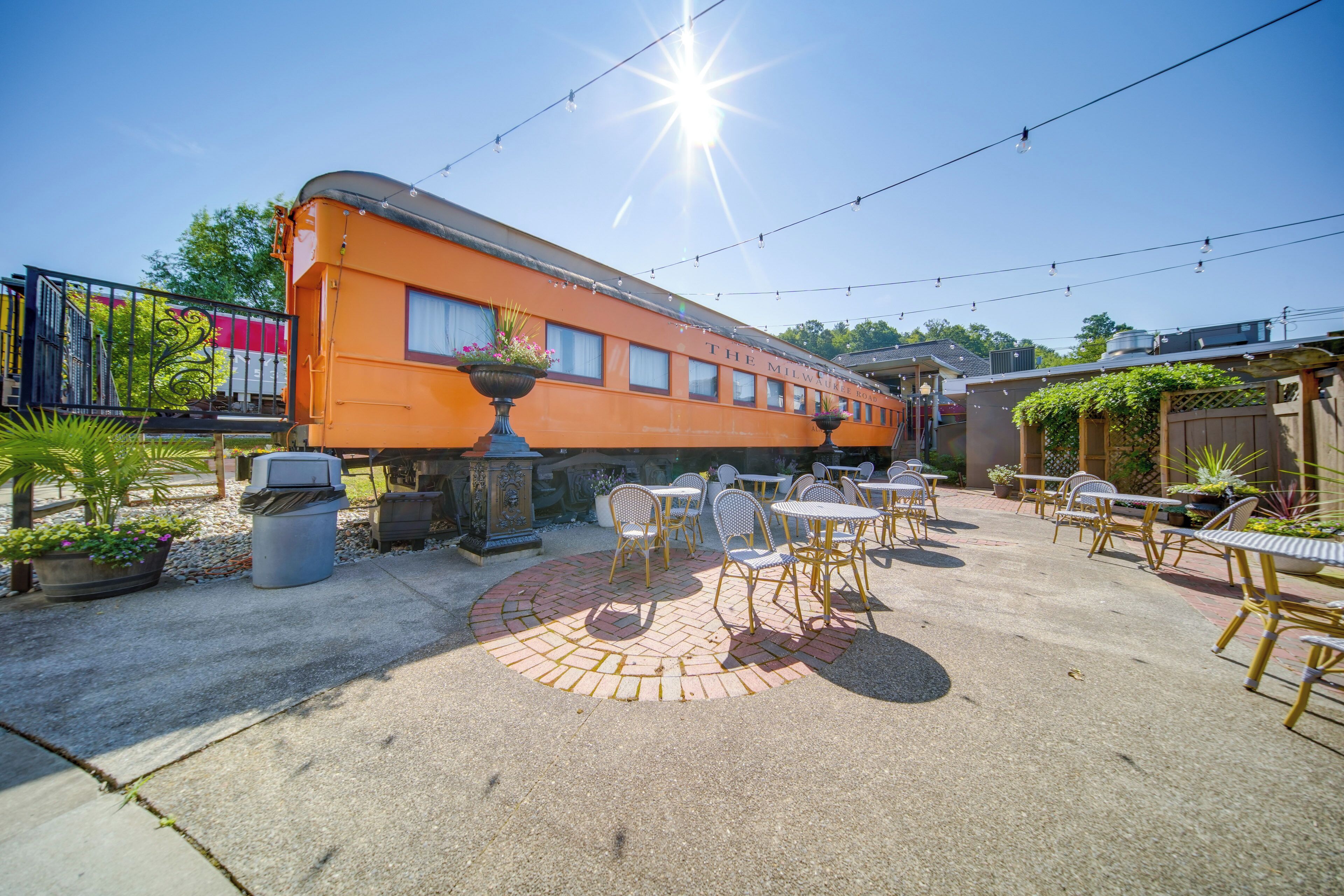 Een terras buiten met tafels en stoelen, een vuurkorf van baksteen en een oud oranje treinwagon.