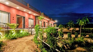 A modern building with a red facade, a covered outdoor area with a swing chair, and a garden with various plants. - Gokul Jawai (Bali)