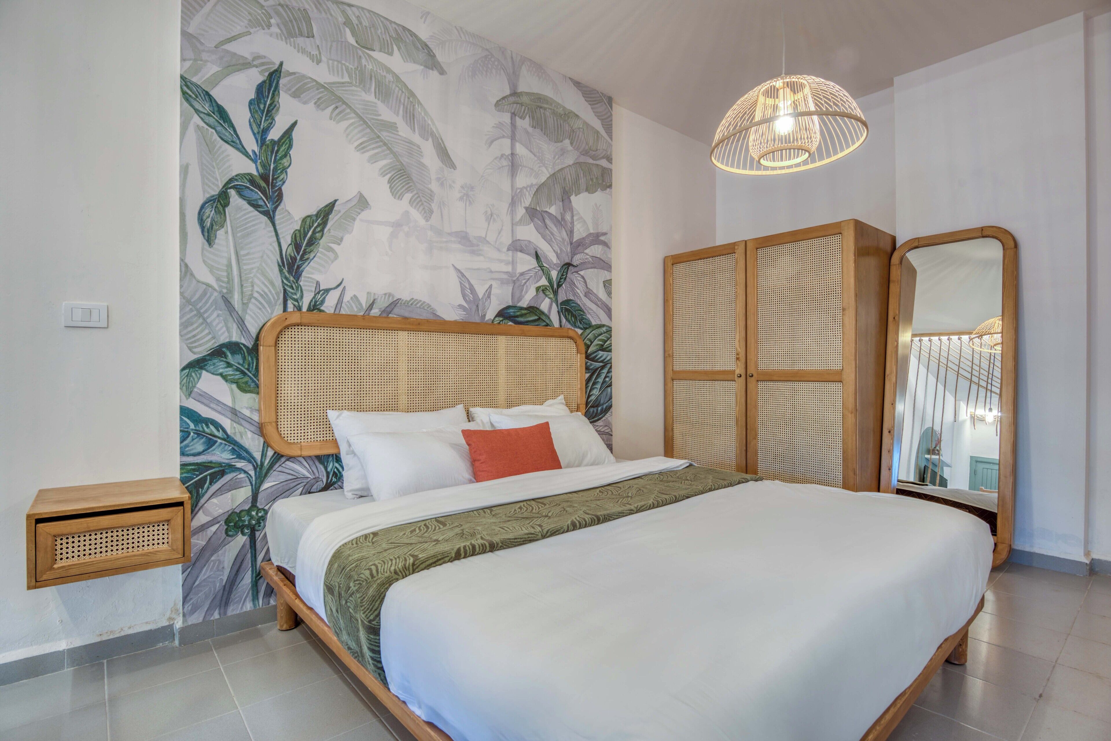 Une chambre avec un grand lit, une tête de lit en rotin, une armoire en osier, un miroir et un luminaire au plafond.