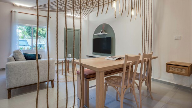A modern living room with a sofa, wooden dining table, and chairs. There is a TV mounted on the wall and a window with curtains. - Natura Studio & Balcony in Batroun (Batroun)