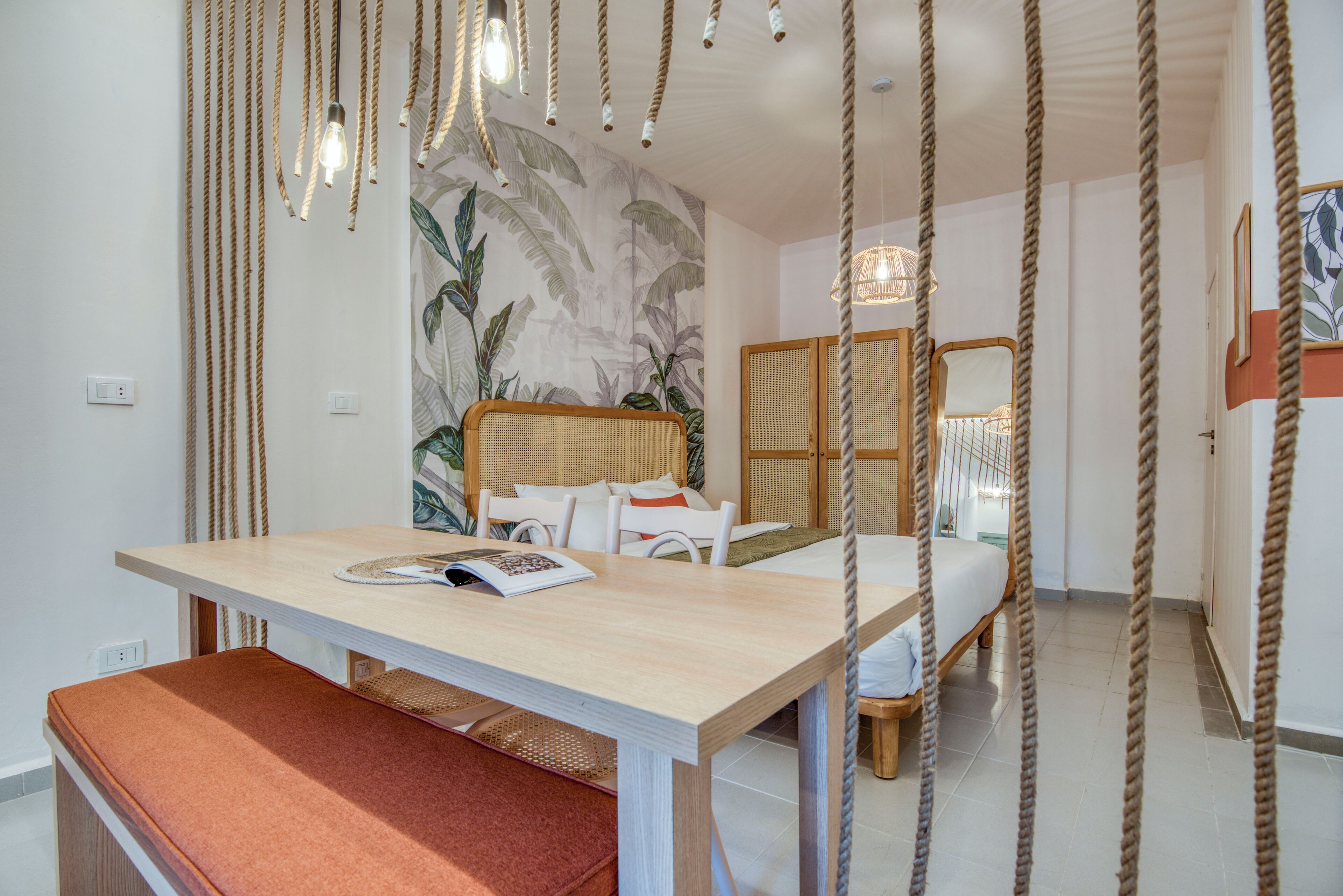 A room with a bed, a bedside table with a book, a bench, and a wall with a tropical mural.