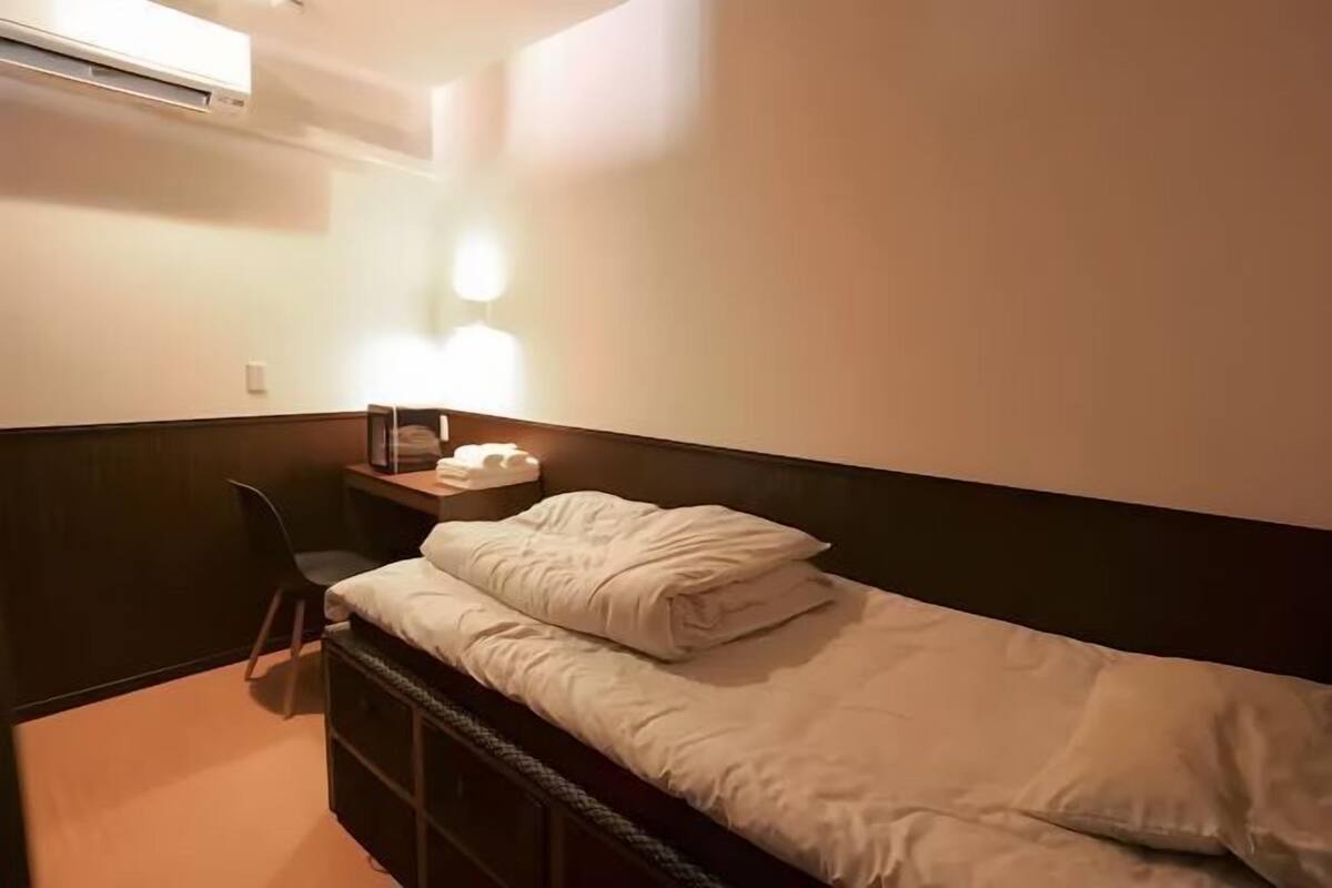 A small, neatly arranged bedroom with a single bed, bedside table, chair, and an air conditioning unit on the wall.