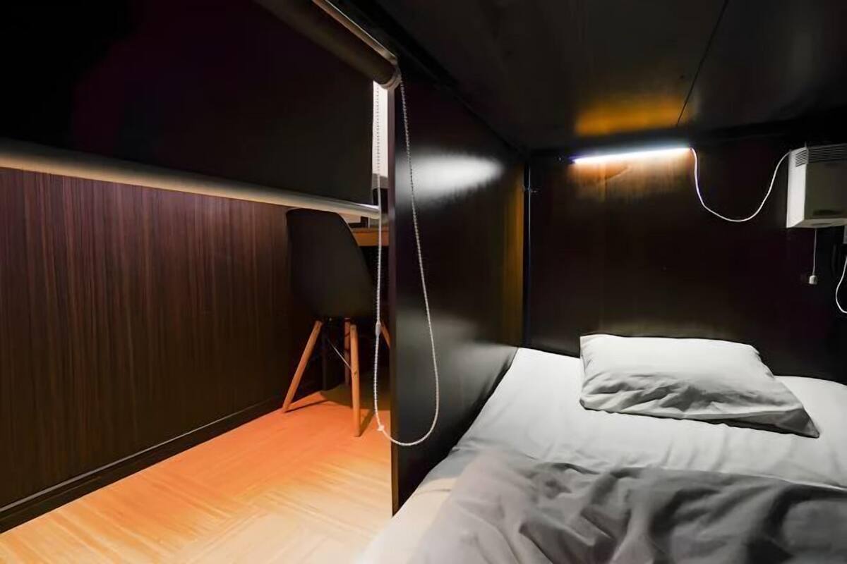 A small, dimly lit room with a single bed, a chair, and a desk.