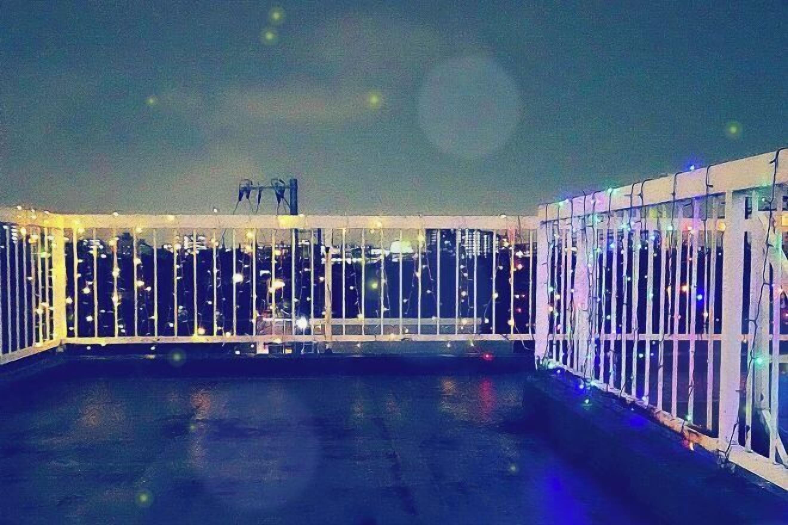 A rooftop terrace with a glass railing, a metal railing, and a view of the city skyline at night.