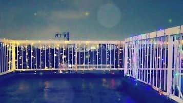 A rooftop terrace with a glass railing, a metal railing, and a view of the city skyline at night.