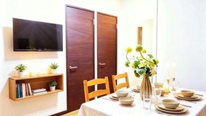 A dining area with a table set for four, a television mounted on the wall, wooden doors, and a bookshelf with books and plants. - Tokyo Yoshimi home stay (Tokyo)