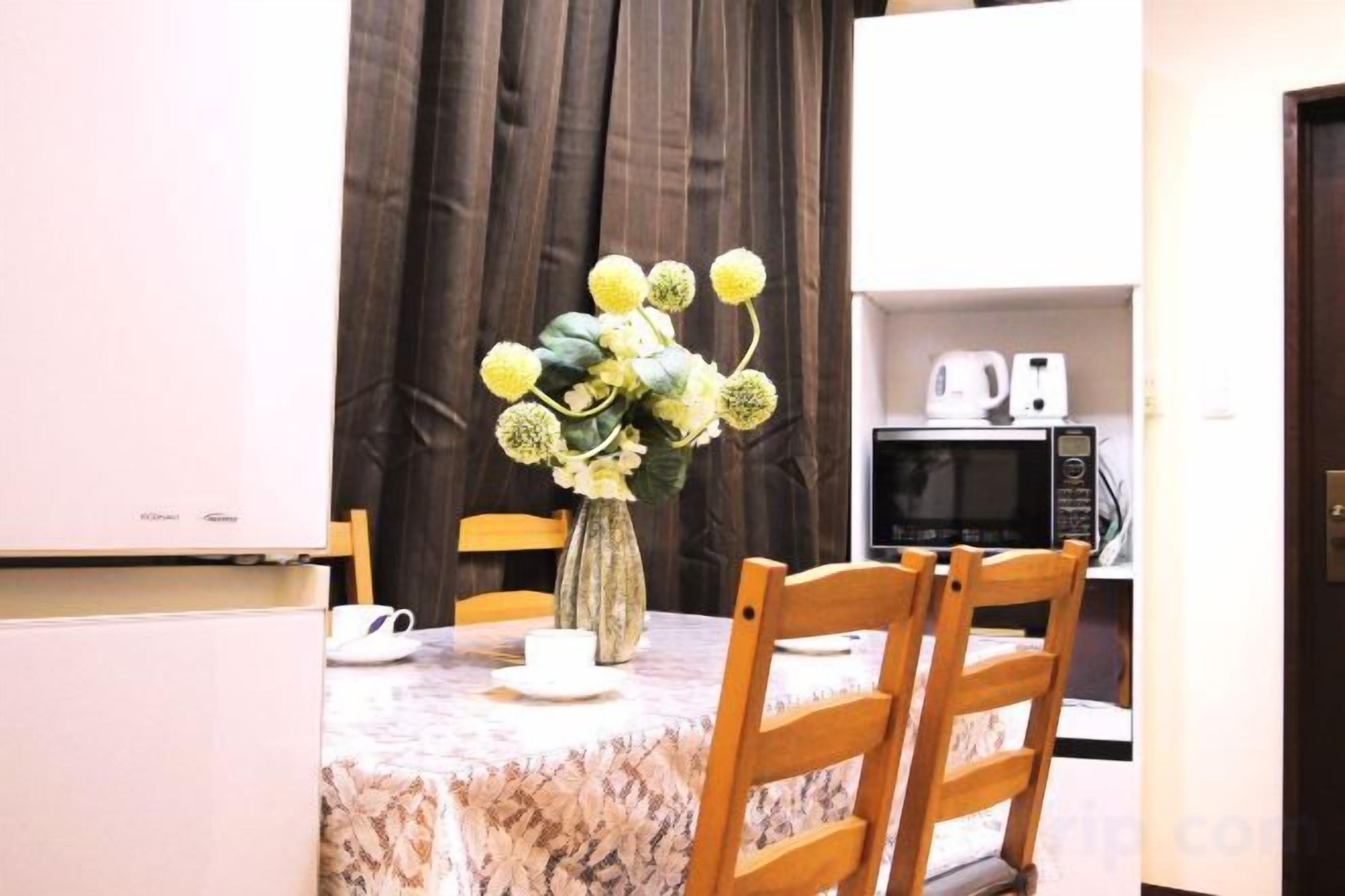 Un comedor con una mesa lista para dos personas, un jarrón con flores y un microondas al fondo.