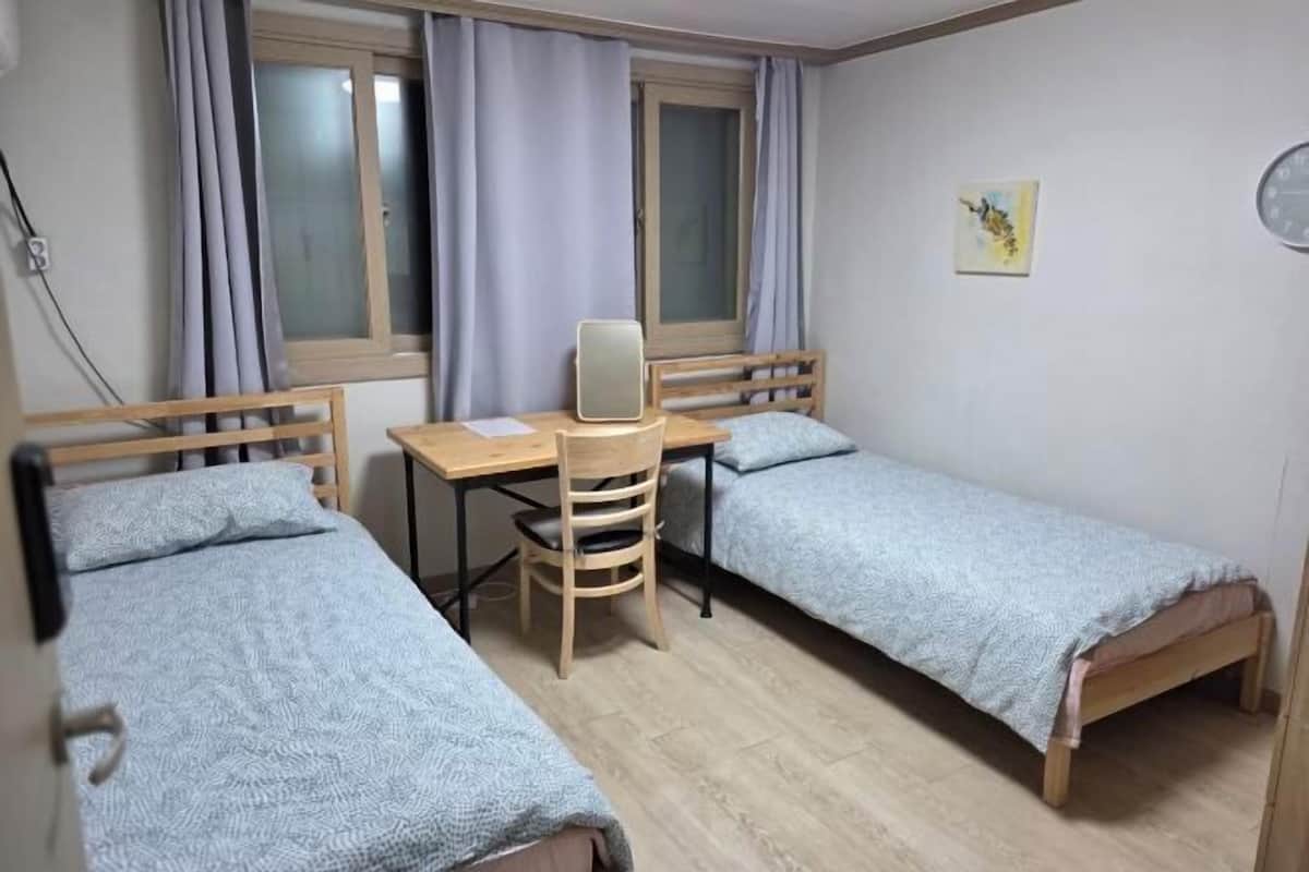 A small, tidy room with two single beds, a wooden desk, a chair, and a window with curtains.